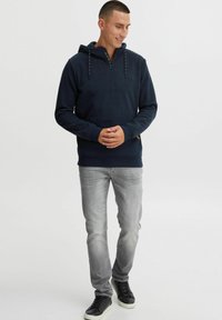 Navy hoodie with a half-zip design, drawstrings, and front pocket, paired with light grey jeans and black sneakers. Smooth texture.