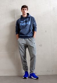 Námornícka mikina s kapucňou a svetlomodrým logom Nike, šedé teplákové nohavice a modré športové topánky. Hladká tkanina, uvoľnený strih a žiadne viditeľné vzory.