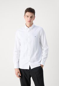 Camicia bianca con maniche lunghe e colletto, con un piccolo logo sul petto, abbinata a pantaloni neri.