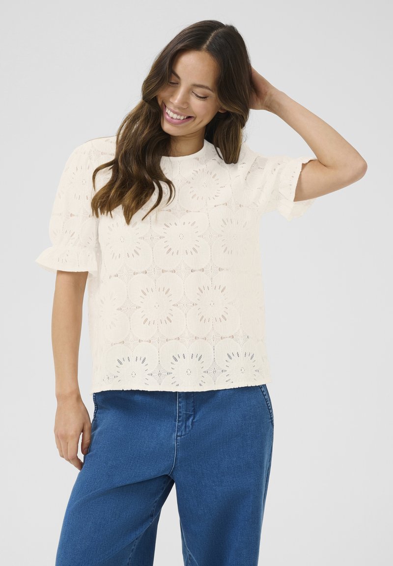 Haut en dentelle crème avec broderie florale, manches courtes et encolure ronde, associé à un pantalon en denim bleu décontracté, mettant en avant un style décontracté.