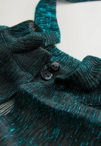 Țesătură tricotată neagră și teal cu fir metalic, având un model cu fante, margini rulate și două butoane negre aproape de decolteu.