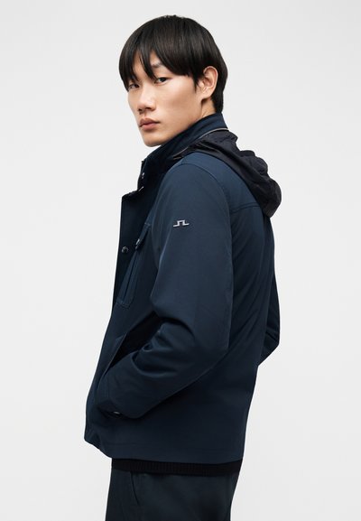 Νεαρός με μαύρα μαλλιά, φορώντας σκούρο μπλε μπουφάν με ανασηκωμένο γιακά και τα χέρια στις τσέπες, κοιτάζοντας πάνω από τον ώμο του.