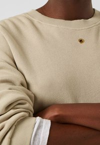 Sweat-shirt beige avec un col côtelé, présentant un petit œillet doré. Tissu texturé et poignets retroussés sur une chemise blanche.