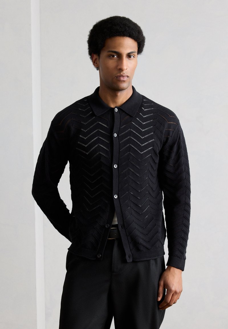 Hombre de piel morena clara y cabello rizado que lleva un suéter negro abotonado con patrones en forma de chevron y pantalones negros, sobre un fondo claro y liso.