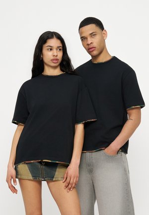 Dvaja modeli stoja blízko seba, oblečení v čiernych veľkých tričkách s maskáčovým lemom, rifľovej mini sukni a svetlosivých nohaviciach, na bielom pozadí.