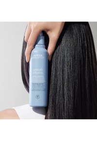 Aveda Smooth Infusion Perfect Blow Dry styling spray i en ljusblå flaska, hålls mot slätt, rakt svart hår. Har vit text och logotyp.