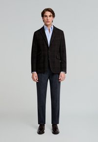 Blazer de cuadros marrones sobre una camisa azul clara, combinado con pantalones gris oscuro y zapatos marrones. Modelo de pie contra un fondo liso.