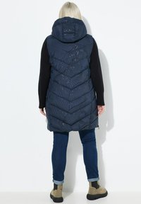 Kvinna med blont hår iklädd en marinblå huva lång vadderad väst, svarta långärmade tröja, blå uppkavlade jeans och beige stövlar, stående vänd mot en vit vägg.