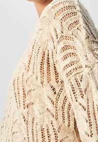 Neulepaita vaaleassa beessissä, jossa on reikämäinen kuvio; ominaisuuksina löysä kudonta lehtimäisellä muotoilulla, korostaen rakennetta ja läpinäkyvyyttä.