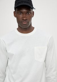 Balta ilgomis rankovėmis palaidinė su apvaliu iškirpimu ir kairiuoju krūtų kišenė; lygus audinys ir minimalus dizainas.