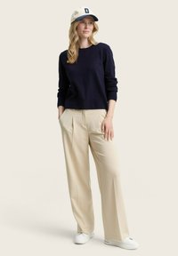 Marine gebreide trui, beige geplooide broek en witte sneakers. Model draagt een crèmekleurige baseballpet met een blauwe letter D. Eenvoudig ontwerp.