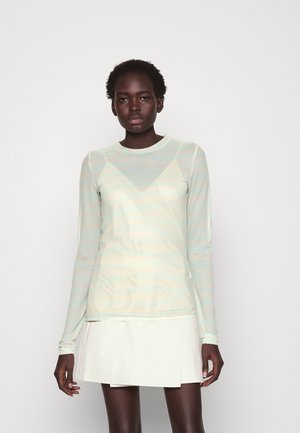 Donkere vrouw in een doorschijnende pastelgekleurde top met lange mouwen over een lichte camisole, gecombineerd met een witte minirok, staande tegen een effen achtergrond.