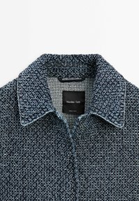 Gros plan sur le col d'une veste bleue en mélange de laine texturée avec une étiquette noire portant la mention Massimo Dutti, fabriquée en Turquie.
