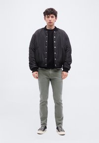 Νέος άντρας στέκεται με το πρόσωπο προς τα εμπρός, φορώντας μαύρο μπουφάν τύπου bomber με κουμπιά, μαύρο πουκάμισο, γκρι παντελόνι και μαύρα-λευκά αθλητικά παπούτσια πάνω σε απλό φόντο.