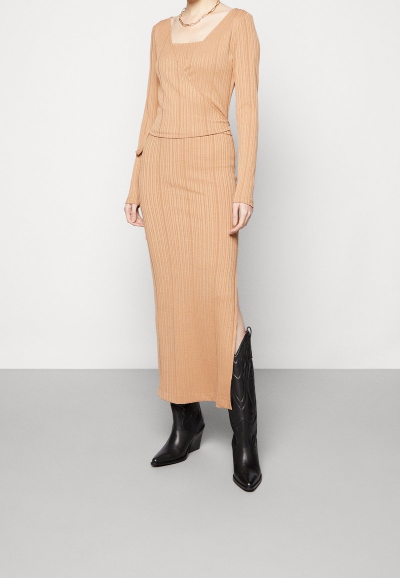 Femme portant un haut beige en maille côtelée à manches longues et une jupe midi assortie avec une fente latérale, associée à des bottes noires à bout pointu mi-mollet.