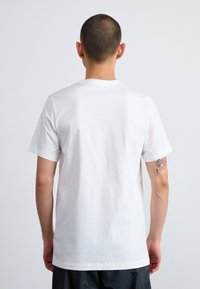 White cotton t-shirt with short sleeves, round neckline, and a plain design, shown from the back. Features visible stitching and a tattoo on the arm.