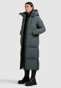 Abrigo acolchado largo en verde oscuro, con capucha, cierre de cremallera, bolsillos laterales y una textura suave. Combinado con botas negras de cordones.