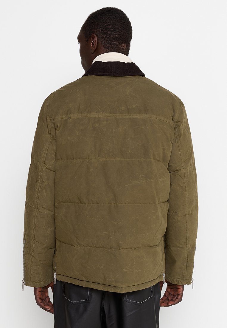 Oliwkowa kurtka puchowa z fakturowanym wykończeniem, zapinanymi na zamek bocznymi kieszeniami i ciemnym kołnierzem. Posiada pikowaną konstrukcję zapewniającą ciepło.
