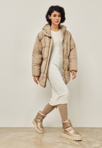 Chaqueta acolchada beige con capucha, cierre de cremallera y textura acolchada; vestido blanco ajustado de canalé; botas beige hasta la rodilla con suela gruesa.