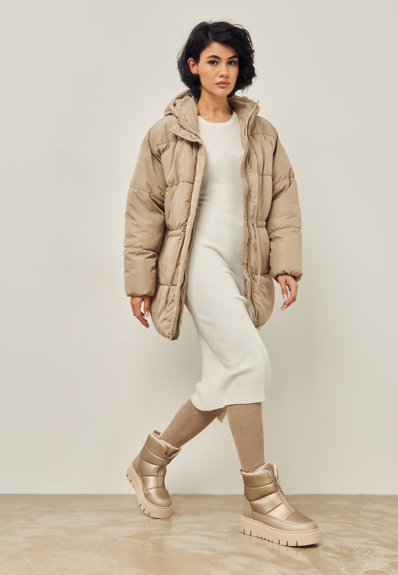 Chaqueta acolchada beige con capucha, cierre de cremallera y textura acolchada; vestido blanco ajustado de canalé; botas beige hasta la rodilla con suela gruesa.