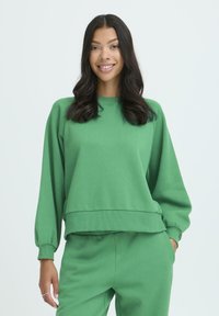 Zelená mikina s dlhými nafúknutými rukávmi, rebrovanými manžetami a lemom, vyrobená z mäkkého materiálu, skombinovaná so zodpovedajúcimi zelenými nohavicami.