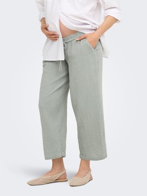 Zwangere persoon draagt lichtgrijze losse broek, eenwit open hemd en beige geweven platte schoenen, staand met één hand in de zak.