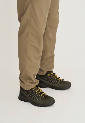 Persoon die beige broek en donkergroene wandelschoenen met gele veters draagt, staand op een effen lichte achtergrond.