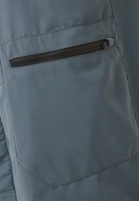 Gros plan sur un tissu bleu avec une poche zippée noire horizontale, montrant des coutures détaillées et la texture.
