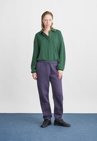 Zeleno rebrast polo majico z dolgimi rokavi in ovratnikom ter tremi gumbi, v kombinaciji z ohlapnimi vijoličastimi trenirkami in črnimi čevlji.