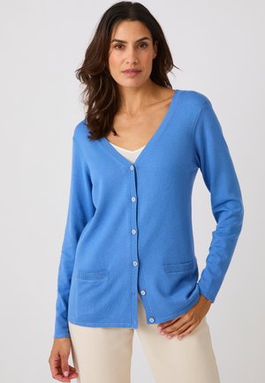 Vrouw met donker haar die een lichtblauwe cardigan met knopen draagt over een wit topje en beige broek, staand tegen een effen lichte achtergrond.