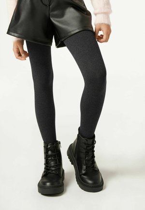 Nohy v černých kožených šortkách, tmavě šedých silonkách a černých šněrovacích kotníkových botách stojící na jednoduchém světlém pozadí.