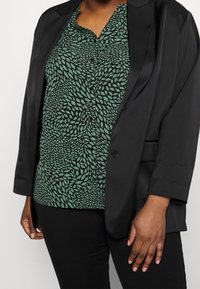 Blazer nero con finitura satinata, abbinato a una blusa verde con motivo a foglie su sfondo nero. Pantaloni neri attillati.