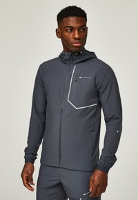 Homme portant une veste à capuche gris foncé et un pantalon assorti avec des poches zippées, debout et regardant de côté sur un fond uni.