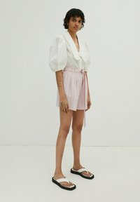 Weiße Bluse mit Puffärmeln und Rüschenausschnitt, kombiniert mit rosa gestreiften Shorts mit einer Krawatte in der Taille, getragen mit weißen Sandalen auf einer schwarzen Sohle.