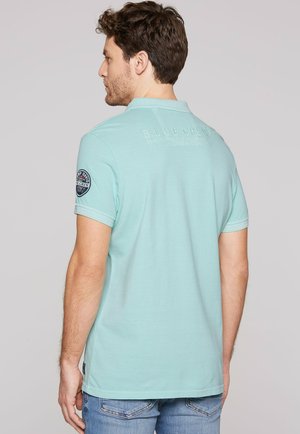 Man met de rug naar de camera, draagt een lichtblauw poloshirt met een patch op de linker mouw en een blauwe spijkerbroek tegen een effen achtergrond.