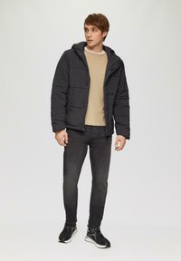 Schwarze Daunenjacke mit Kapuze, ausgestattet mit einem gesteppten Design und vorderen Taschen, getragen über einem beigen Pullover und schwarzen Jeans mit Turnschuhen.
