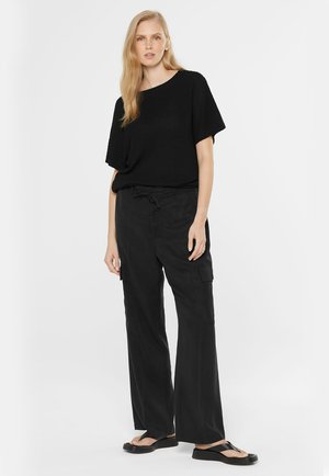 Frau steht und trägt ein lockeres, schwarzes, kurzärmliges Strickoberteil, breite schwarze Cargohosen und schwarze Plateausandalen vor weißem Hintergrund.