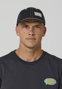 Fekete sapka, varrott karimával és logófolttal. Sötét póló felett viselve, színes márkajelzéssel. Semleges háttér.