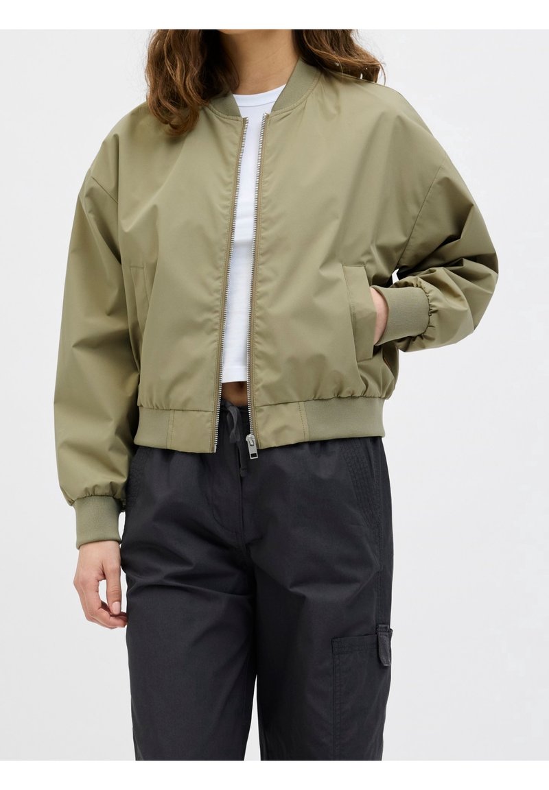 Personne portant une veste bomber vert olive sur un crop top blanc et un pantalon cargo noir, avec une main dans la poche.