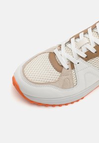 Baskets beige et blanches à l'avant avec une tige en mesh, des superpositions en cuir et en suède, des lacets blancs et des détails orange sur la semelle en caoutchouc.
