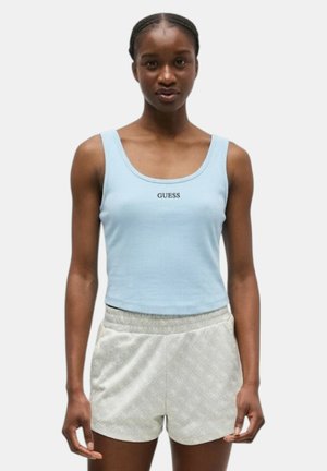 Donna che indossa un top corto senza maniche azzurro chiaro con la scritta "GUESS" e pantaloncini grigio chiaro testurizzati, in piedi contro uno sfondo semplice.
