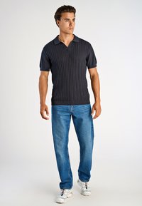 Homme portant un polo en maille bleu marine à manches courtes, un jean bleu et des baskets blanches, debout les mains détendues, regardant sur le côté sur un fond uni.