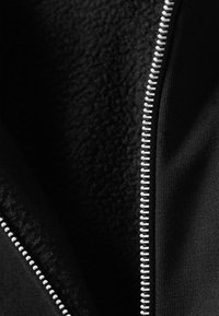 Crni odjevni predmet sa patentnim zatvaračem, unutrašnjosti od flisa i glatke tkanine izvana. Srebrni detalj patentnog zatvarača proteže se duž otvora. Vidljiv je teksturirani podstavljeni sloj.