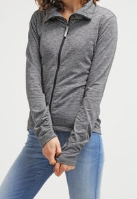 Femme portant une veste grise ajustée à fermeture éclair avec des ouvertures pour les pouces et un jean bleu clair, debout devant un fond uni.