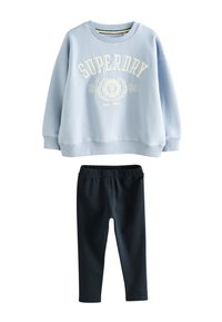 Šviesiai mėlynas medvilninis džemperis su „SUPERDRY“ logotipu ir herbu kreminės spalvos, derinamas su juodos spalvos tamprėmis tamprėmis. Kasdienis, laisvo silueto derinys.