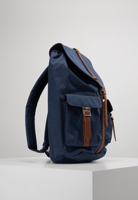 Granatowy plecak z materiału z brązowymi skórzanymi akcentami, z górną klapą, przednią kieszenią oraz regulowanymi, wyściełanymi szelkami.