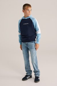 Morský a svetlomodrý farebne blokovaný hoodie s bielymi akcentmi, bavlnený materiál, klokaní vrecko, spárovaný s roztrhnutými modrými džínami a čiernymi teniskami.
