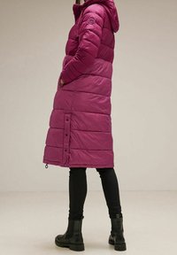 Lang pufferjakke i fuchsia, med horisontale quiltede sektioner, en snoretrækshætte og trykknaplukninger i kanten. Sammen med sorte skinny jeans og støvler.