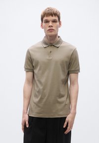 Camisa polo caqui de mangas curtas, ajuste slim, gola de malha lisa e detalhe de logótipo subtil no peito. Textura de tecido suave.