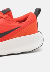 Rød sporty sko med en teksturert mesh-overdel og svarte detaljer. Har en tykk hvit mellomsåle og en teksturert oransje hælpanel.